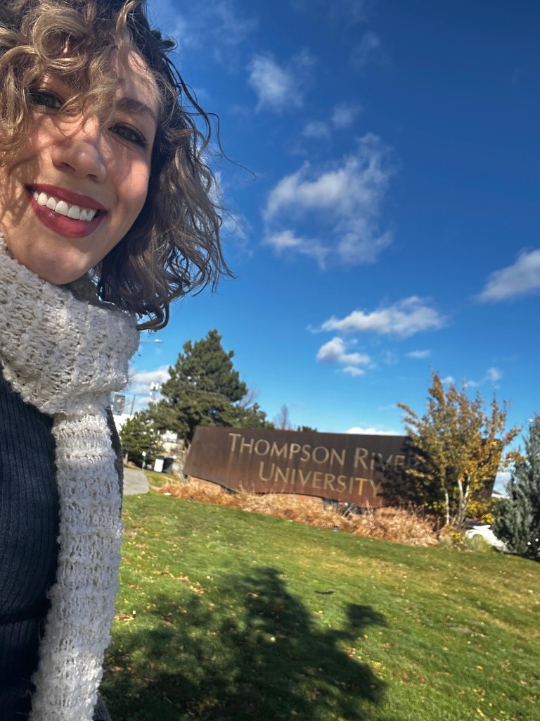Dora María González at Thompson Rivers University, with the TRU entrance sign visible behind her on a grassy hill